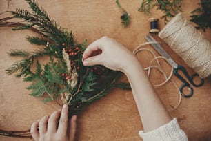 Christmas wreath workshop. Hands holding fir branches, pine cones, berries, thread, scissors on wooden table, flat lay. Making rustic christmas wreath. Authentic rural wreath