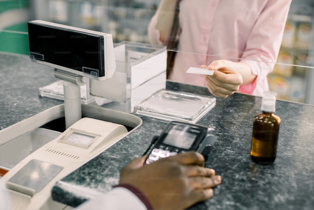 Customer woman making payment in drugstore. Pharmacist holding terminal, woman paying for drugs with credit card. Modern payment machine, syrup on counter