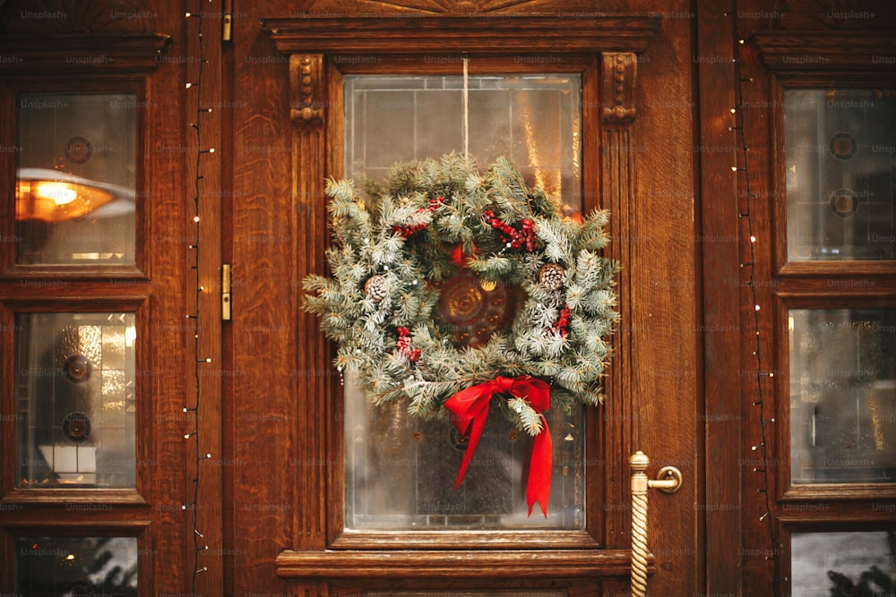 Stylish christmas wreath with red bow and berries at front doors of store at holiday market in city street. Space for text. Rustic decoration. Christmas street decor.
