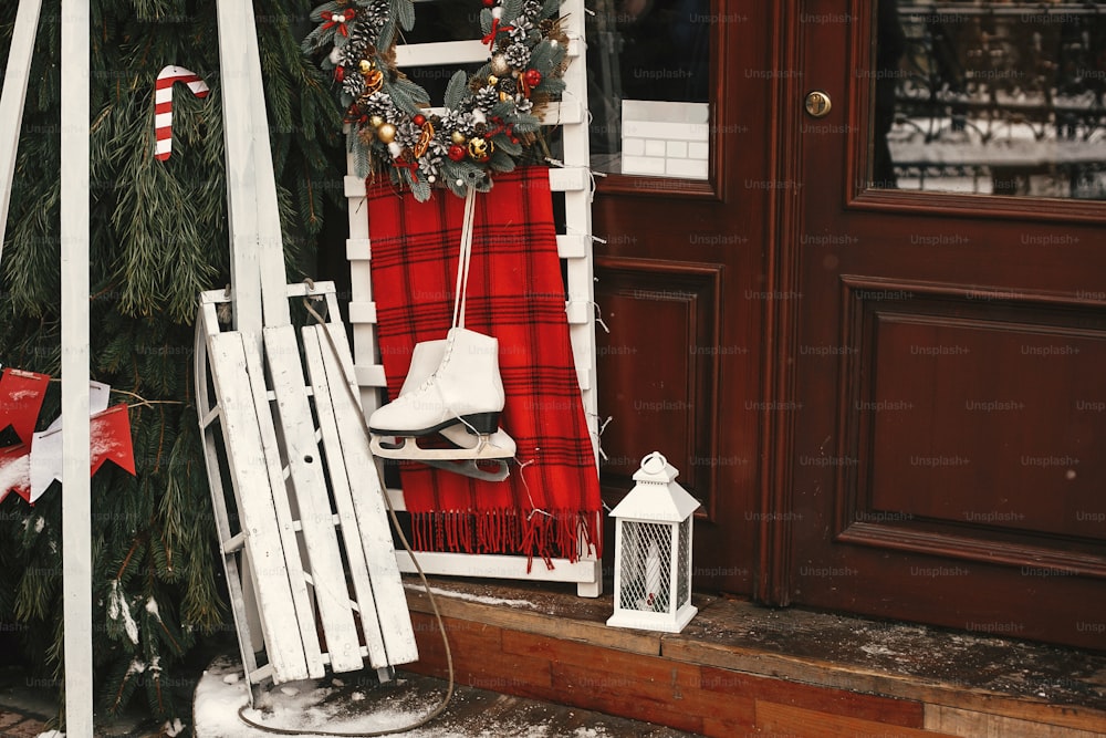 Stylish christmas wreath, white wooden sleigh and ice skate at front of store at holiday market in city street. Christmas street decor. Space for text. Rustic decoration