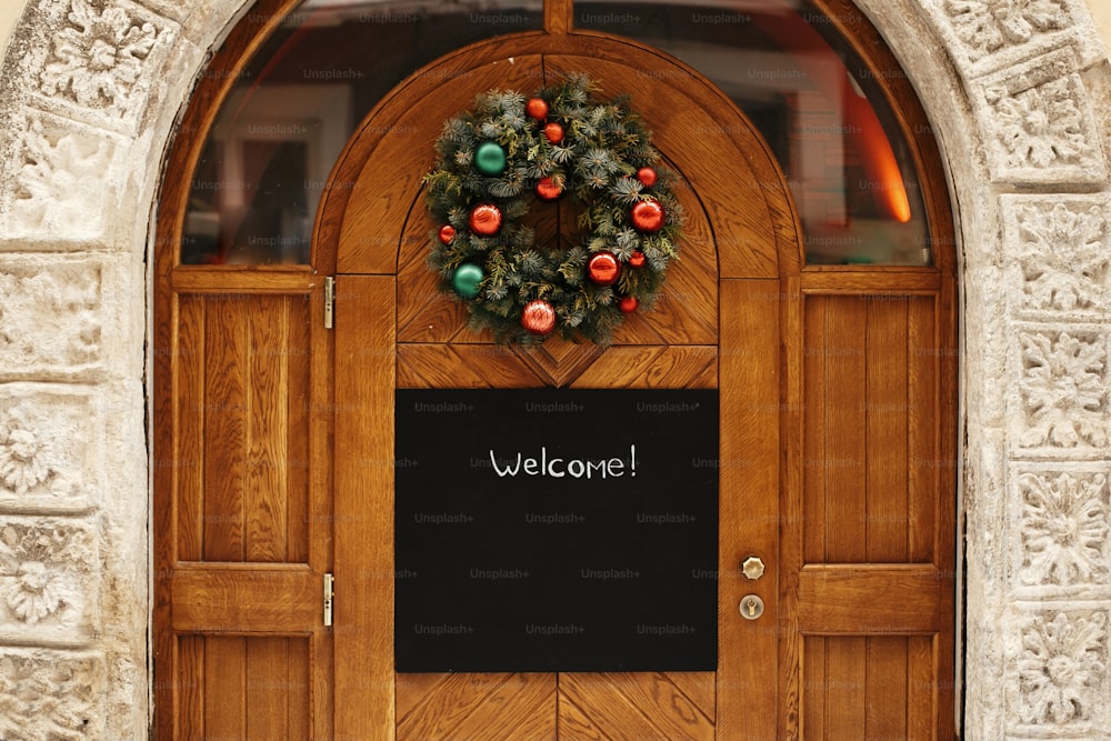Christmas street decor. Stylish christmas wreath with red and green baubles and empty board with copy space on rustic wooden doors at holiday market in city street. Space for text.  template