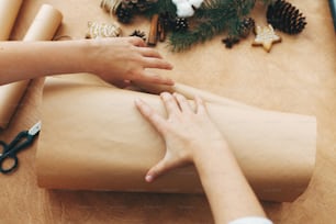 Wrapping christmas gifts concept. Hands wrapping christmas gift in craft paper and pine branches, cones, gingerbread cookies, thread, cinnamon, scissors on rural wooden table