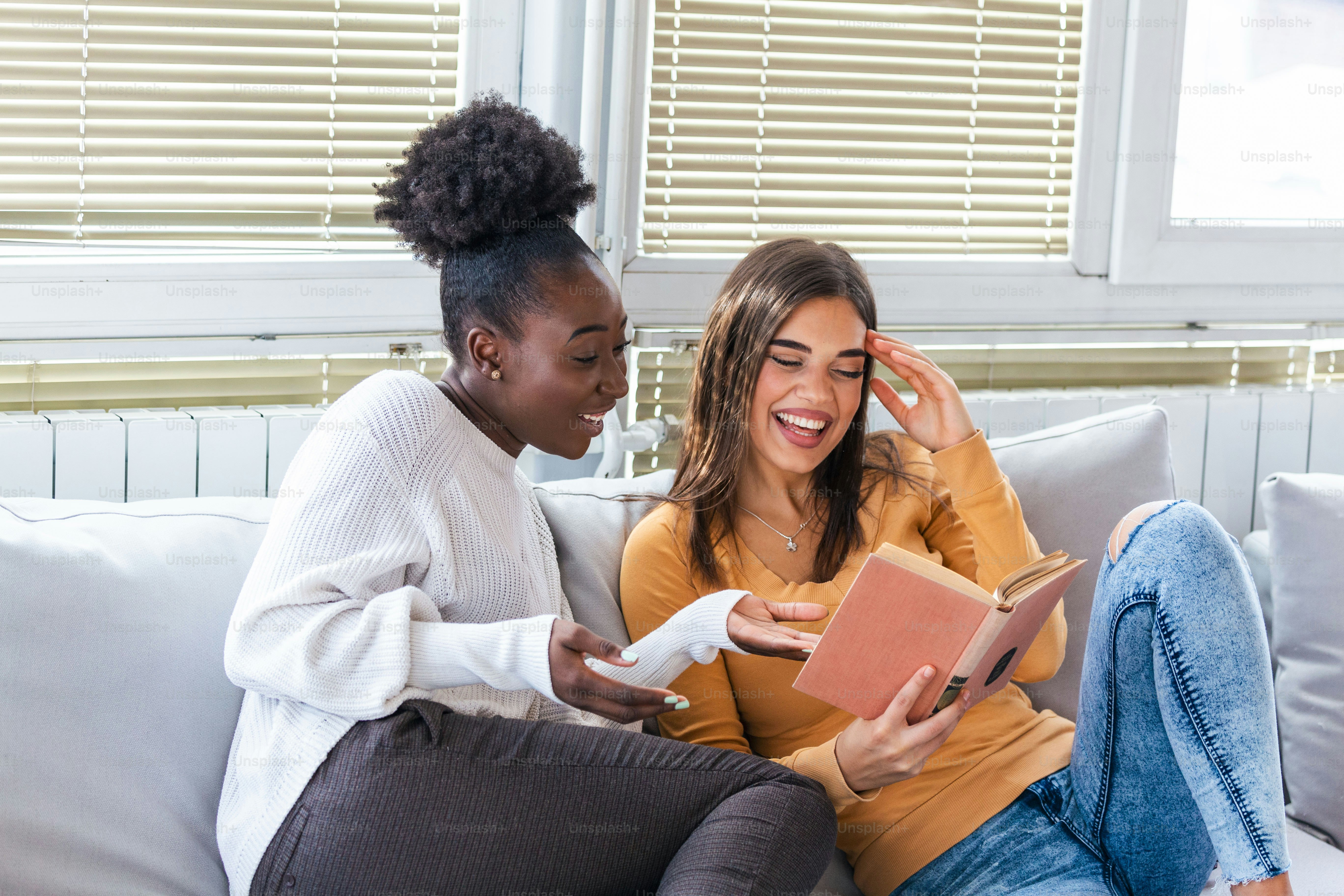 Two women laughing about funny stories from books at home. Friendship and care, happy girl friends. African American and caucasian best friends