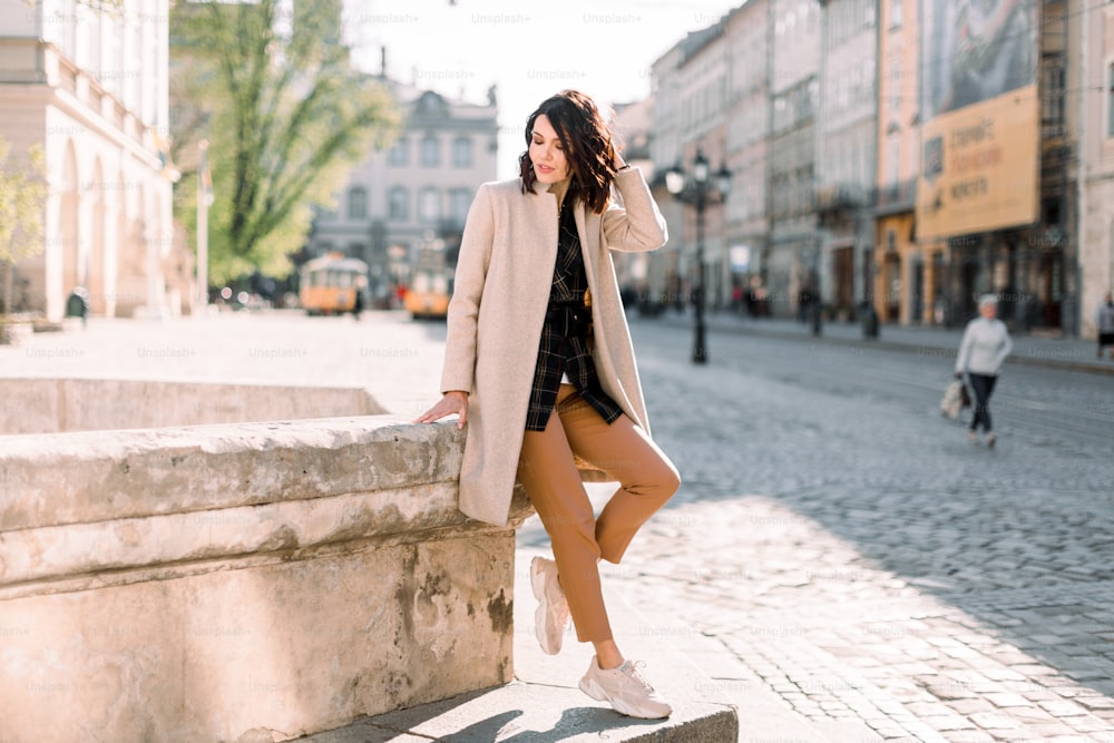 Beautiful fashionable woman in beige coat standing on the old city street. Ancient buildings, morning city background.