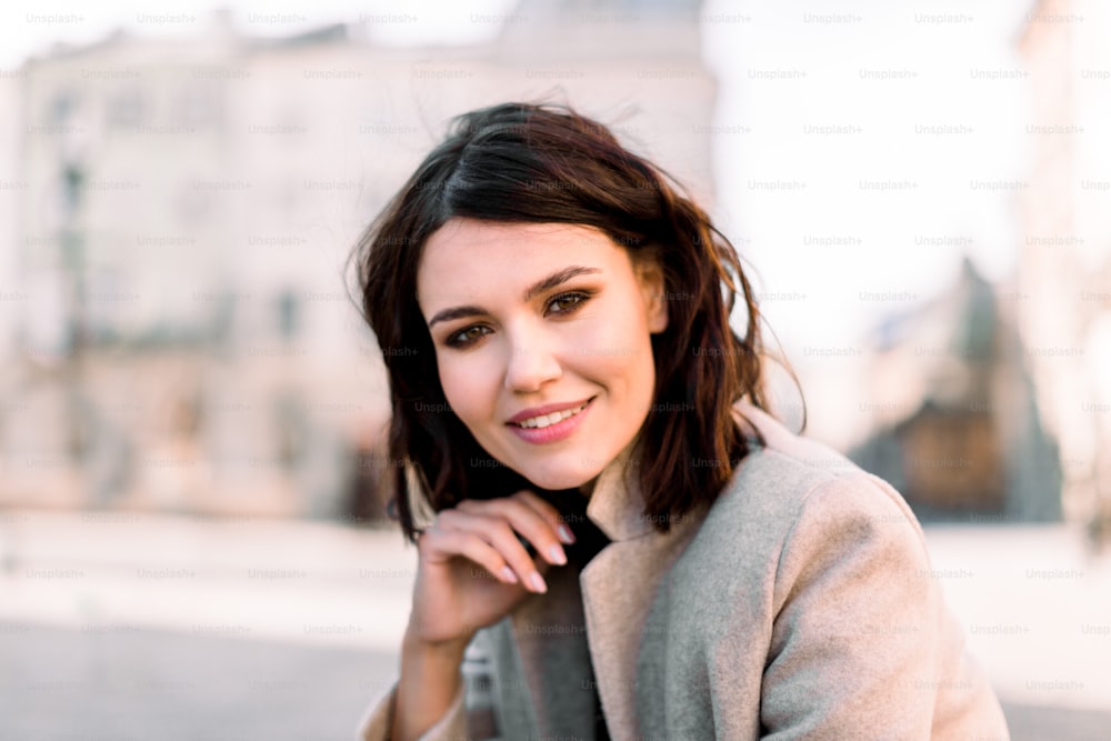 Lovely young woman, wearing in beige coat, with black short hair, posing on the street of old European city, on sunny day, close-up, copy space.