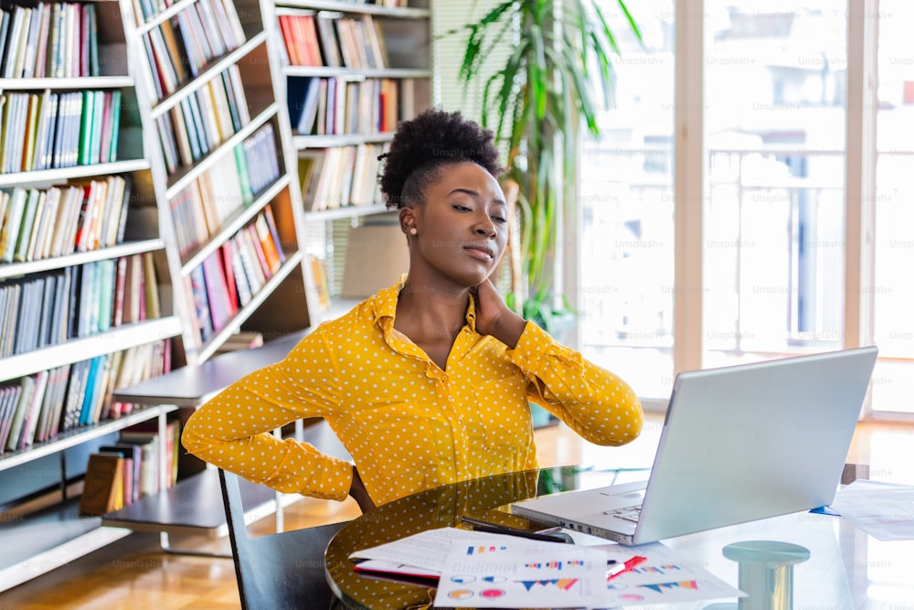 Portrait of young black stressed woman sitting at home office desk in front of laptop, touching aching back with pained expression, suffering from backache after working on pc