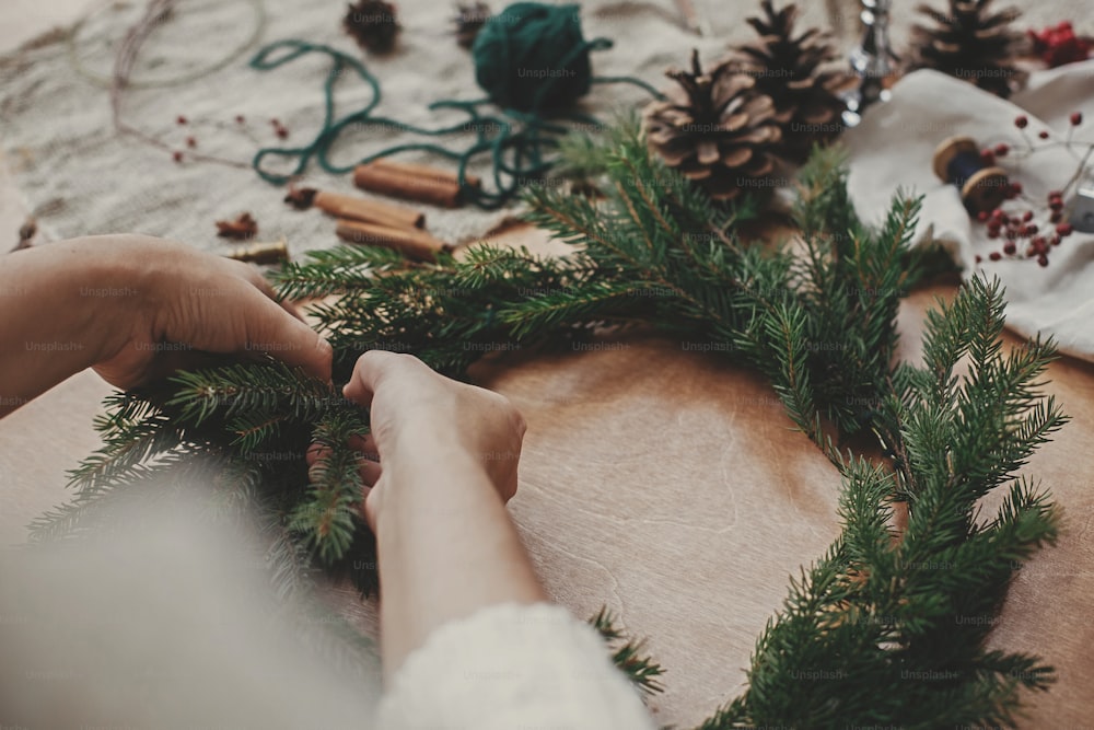 소박한 크리스마스 화환 만들기. 전나무 가지와 소나무 콘, 실, 열매, 나무 테이블에 가위를 들고 손. 크리스마스 화환 워크숍. 정통 세련 된 정물