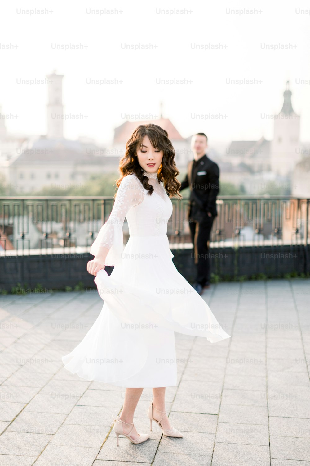 Joven hermosa novia asiática en vestido de novia blanco bailando en la terraza de la ciudad antigua. Novio de pie en el fondo. Paseo nupcial en la ciudad vieja.