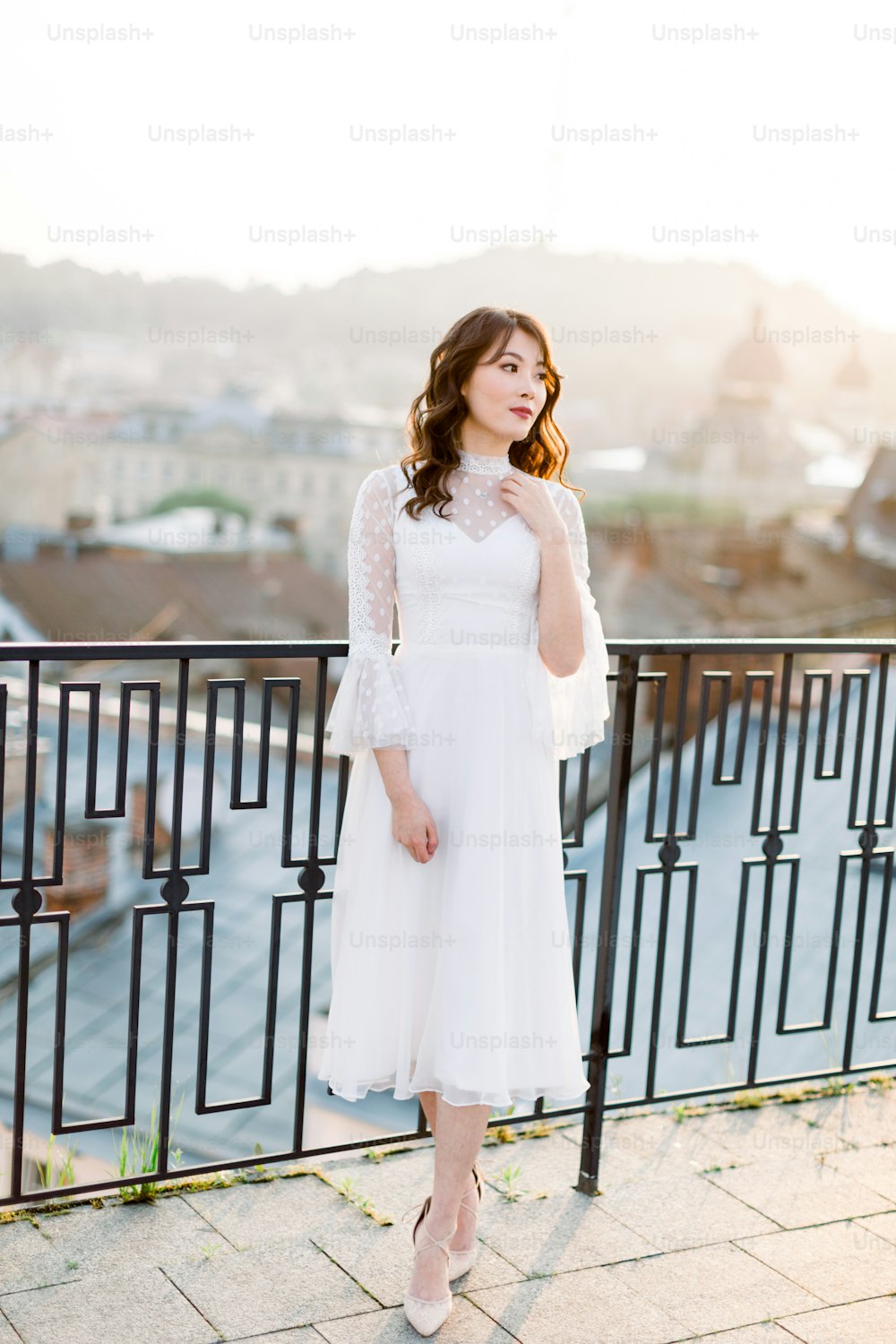 Elegant asian woman in white dress standing on rooftop terrace looking at city background skyline view of old European city in sunset. Chinese bride posing on the background of old city view.