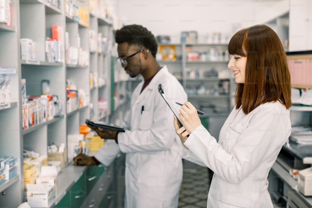 Confident Male And Female Pharmacists In Pharmacy. African American male pharmacist working on digital tablet and Caucasian woman making notes on clipboard during inventory in pharmacy.