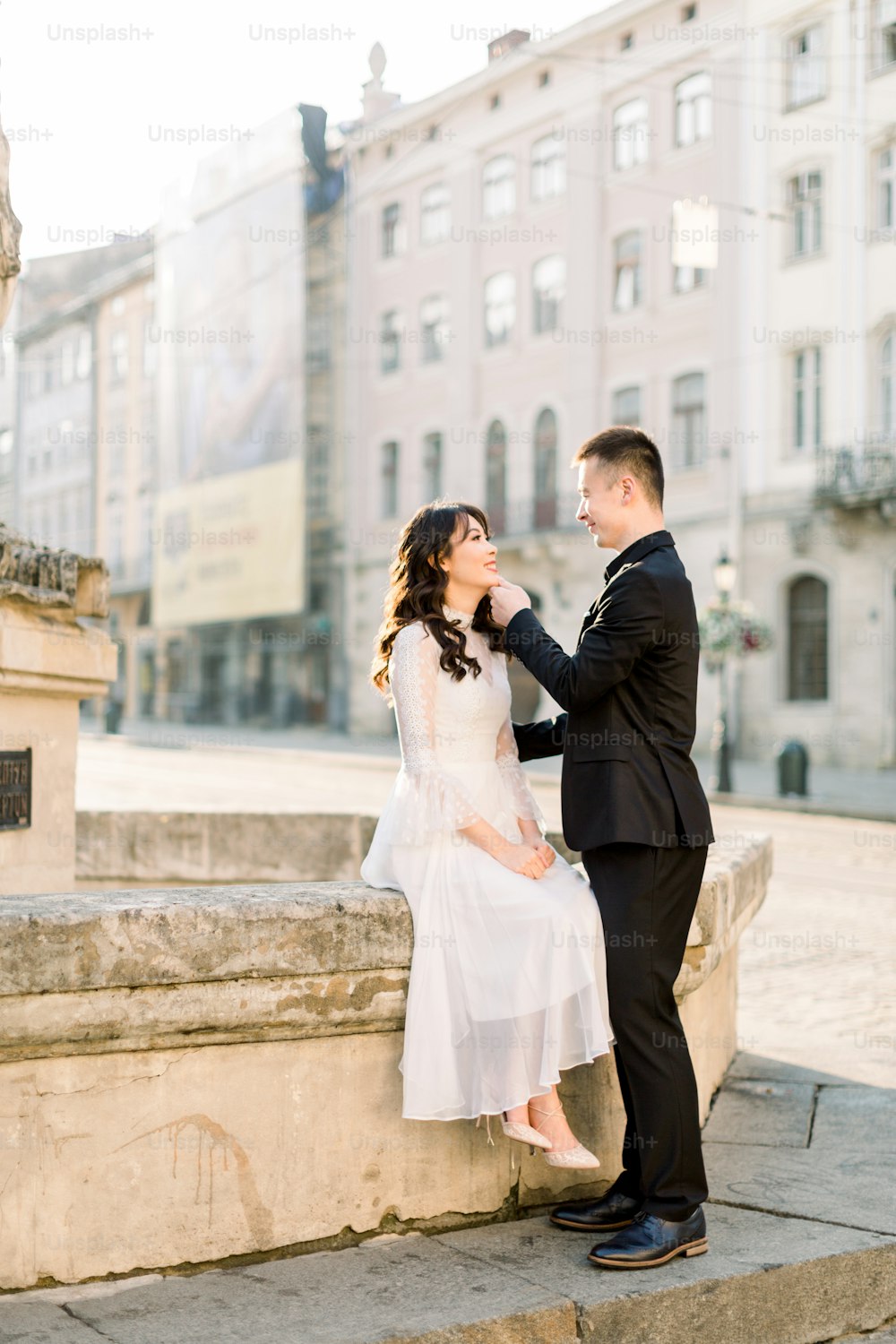 Stylish beautiful couple Asians newlyweds walking on the old city center streets on a sunny day of their wedding. Woman in white dress and man in black suit looking each other.