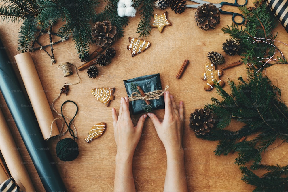 Joyeux Noël, plat rustique. Mains tenant un cadeau de Noël élégant et des branches de pin, des cônes, des biscuits en pain d’épice, du fil, de la cannelle, du coton sur une table en bois rurale. Meilleurs vœux