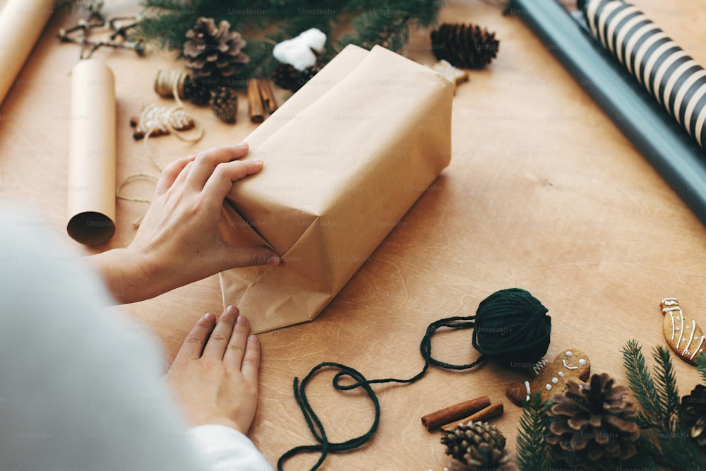 Hände wickeln Weihnachtsgeschenke in Bastelpapier und Tannenzweige, Zapfen, Lebkuchenplätzchen, Faden, Zimt auf ländlichem Holztisch. Stilvolles Weihnachtsgeschenkkonzept verpacken