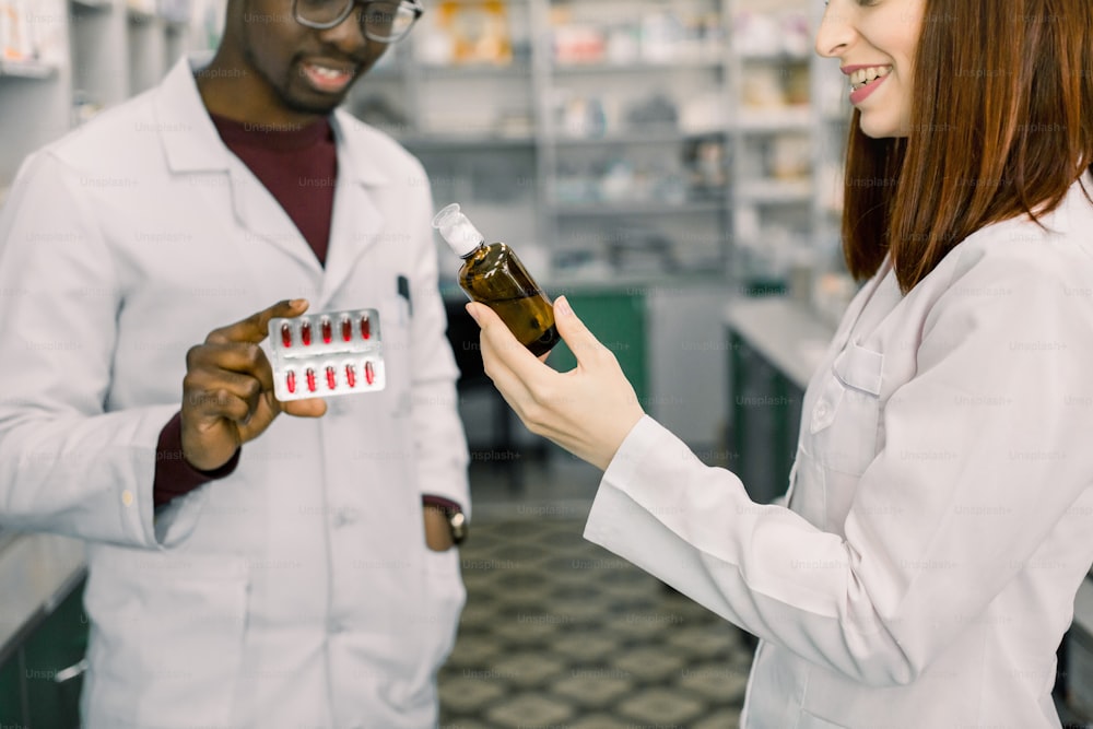 Closeup cropped portrait of two smiling friendly pharmacists working in modern farmacy. African man holds red pills blister and Caucasian woman holds bottle of syrup.