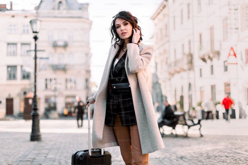 Bella giovane donna con i capelli scuri corti in abiti eleganti casual che viaggia con una valigia a ruote, camminando per la strada della vecchia città europea.