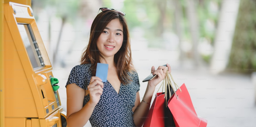Mulher asiática nova segurando o cartão de crédito e saco de compras depois de retirar o dinheiro da máquina ATM