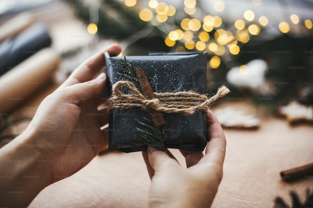 Manos sosteniendo regalo rústico de navidad en papel de regalo negro con decoración de canela sobre fondo de luces doradas, ramas de pino, conos, galletas de jengibre. Feliz Navidad