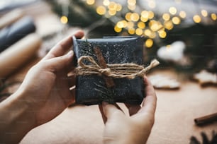 Hands holding rustic christmas gift in black wrapping paper with cinnamon decoration on background of golden lights, pine branches, cones, gingerbread cookies. Merry Christmas