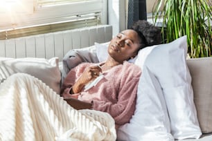 Krankheit, saisonales Virusproblemkonzept. Frau, die krank ist und Grippe hat, liegt auf dem Sofa und schaut auf die Temperatur auf dem Thermometer. Kranke Frau liegt mit hohem Fieber im Bett. Erkältungsgrippe und Migräne.