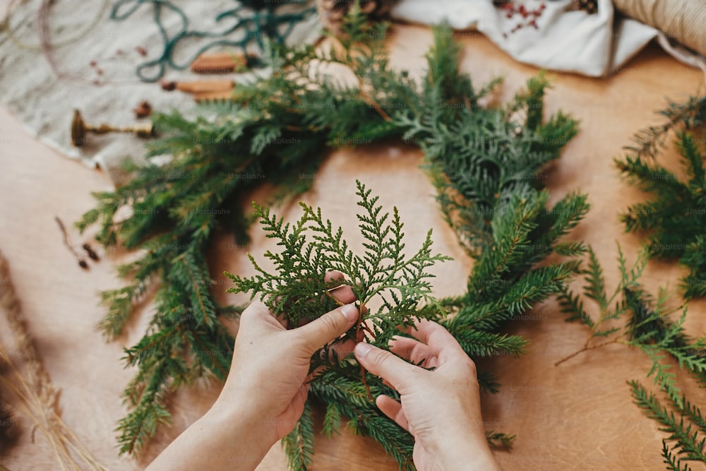 Hands holding fir branches, and pine cones, thread, berries, scissors on wooden table. Christmas wreath workshop. Authentic stylish still life. Making rustic Christmas wreath.