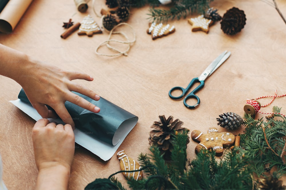 Emballage de cadeaux de Noël concept. Mains emballant le cadeau de Noël dans du papier noir et des branches de pin, des cônes, des biscuits en pain d’épice, du fil, de la cannelle, des ciseaux sur une table en bois rurale