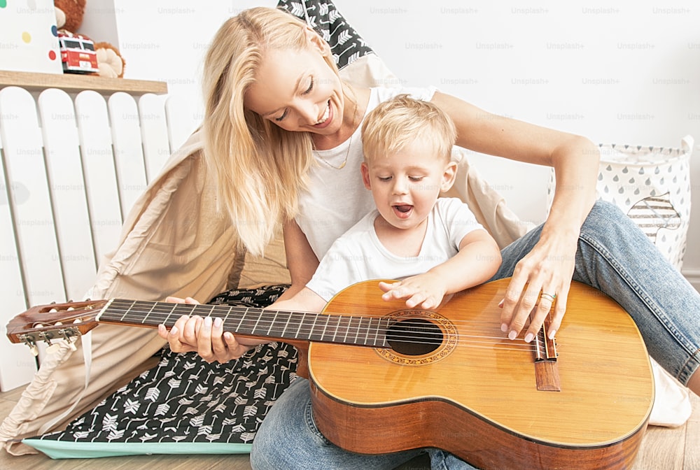 Mother and little child playing in kids room. Mom with son making music on guitar. Leisure time at home. Family happiness and love.