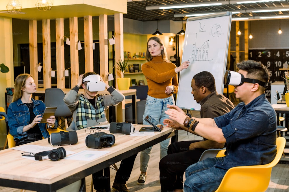 Young woman leader of the project speaking to her colleagues standing near the whote office board. Multiracial team of people having fun wearing virtual reality glasses in office sitting at the table.