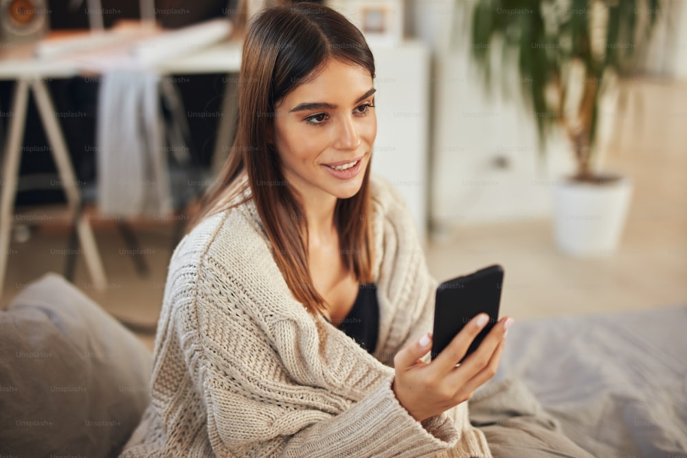 Attractive caucasian brunette dressed in beige sweater sitting on bed in bedroom and using smart phone.