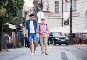Un hombre joven y un anciano ciego con bastón blanco caminando por la acera en la ciudad.