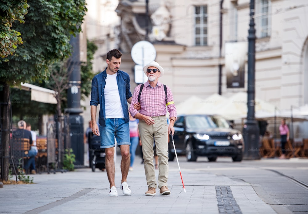 A young man and blind senior with white cane walking on pavement in city.