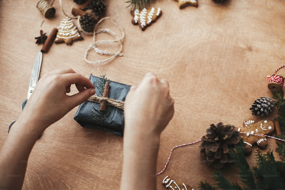 Regalo di Natale rustico da incartare. Mani che confezionano il regalo di Natale in elegante carta nera e rami di pino, coni, biscotti di pan di zenzero, filo, cannella su tavolo di legno rurale.
