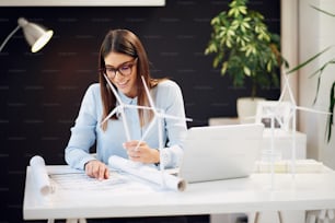 Charming hardworking brunette dressed in formal wear sitting at desk in office, holding windmill model and looking at plans. On desk is laptop.