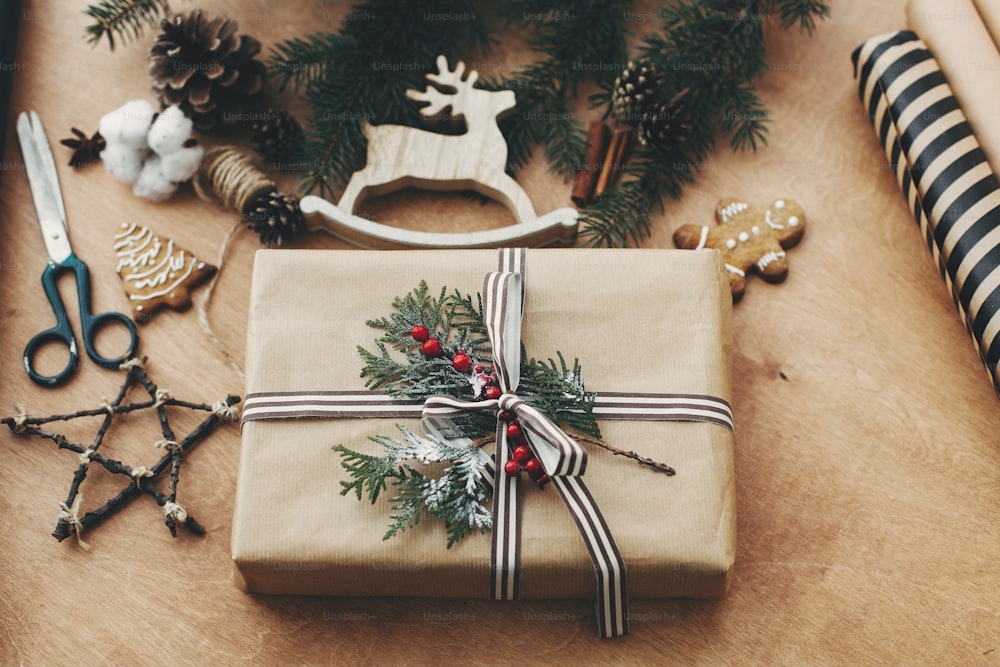 세련된 소박한 크리스마스 선물 상자, 포장지, 나무 순록, 소나무 가지와 콘, 시골 나무 테이블에 진저 브레드 쿠키. 즐거운 성탄절. 계절 인사말 카드