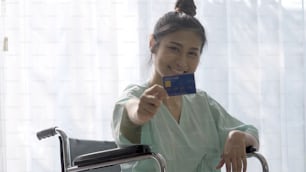 Glücklicher Patient hält Kreditkarte und l�ächelt, während er nach der Genesung von einer Verletzung auf der Krankenhausstation im Rollstuhl sitzt. Medizinische Versorgung und gutes Patientengeld-Zahlungsservice-Konzept.