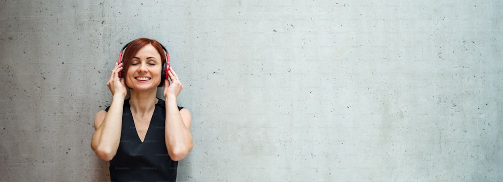 A portrait of young business woman with headphones standing against concrete wall in office and listening to music.