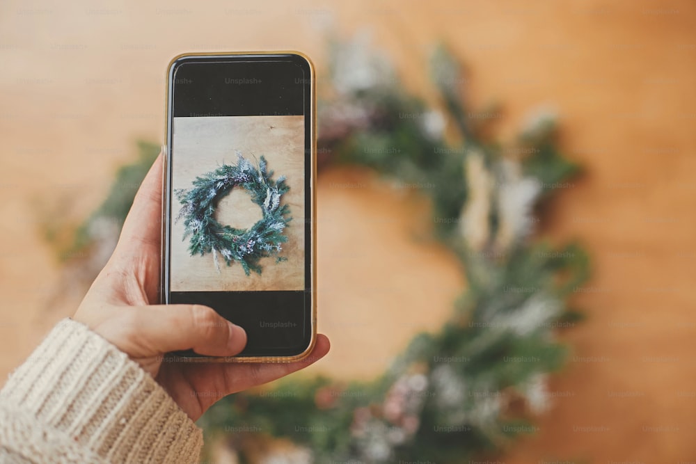 Hand holding phone and taking photo of rustic Christmas wreath flat lay. Christmas mockup. Hipster girl making photo of christmas wreath on smartphone. Merry Christmas