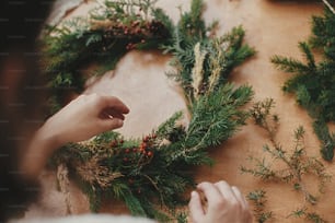 Making rustic Christmas wreath. Hands holding fir branches, and pine cones, thread, berries on wooden table. Christmas wreath workshop. Authentic stylish still life