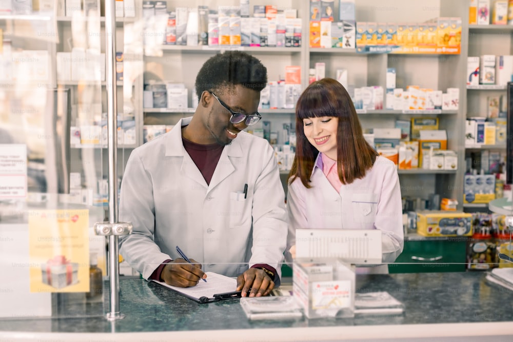 Confident multiethnical Male And Female Pharmacists In Pharmacy. African American man pharmacist making notes on clipboard during inventory in pharmacy.