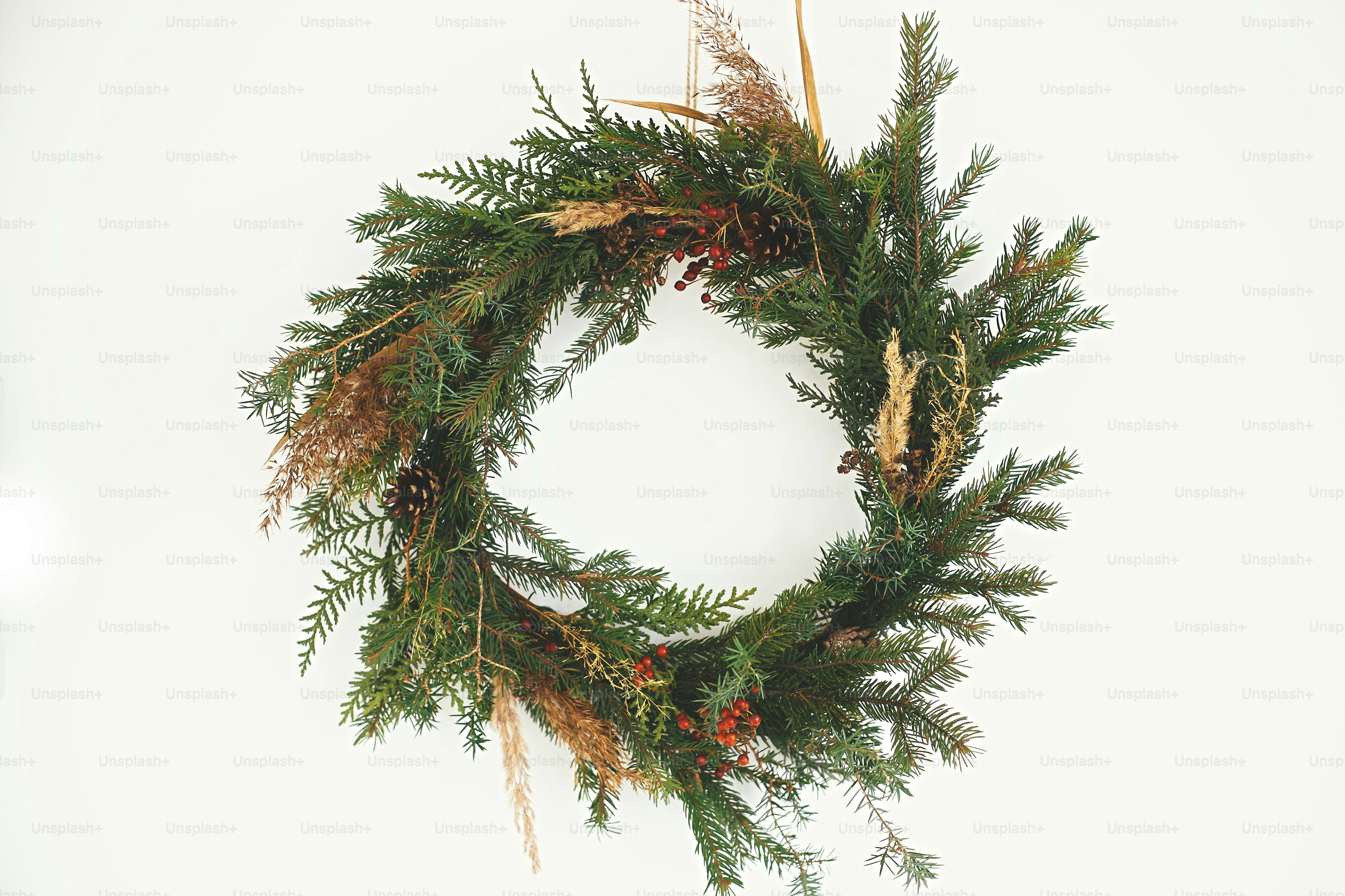 Corona rústica de Navidad. Corona navideña rural creativa con ramas de abeto, bayas, piñas y hierbas que cuelgan en la pared blanca de la habitación. Espacio de copia. Saludos de temporada.