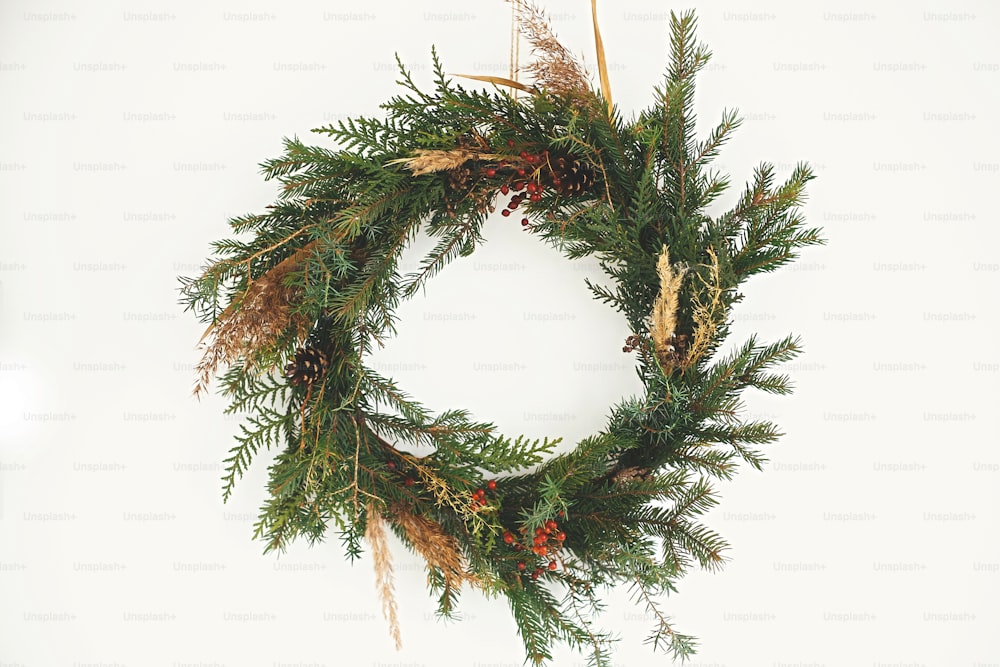 Christmas rustic wreath. Creative rural christmas wreath with fir branches, berries, pine cones and herbs hanging on white wall in room. Copy space. Season's greetings.