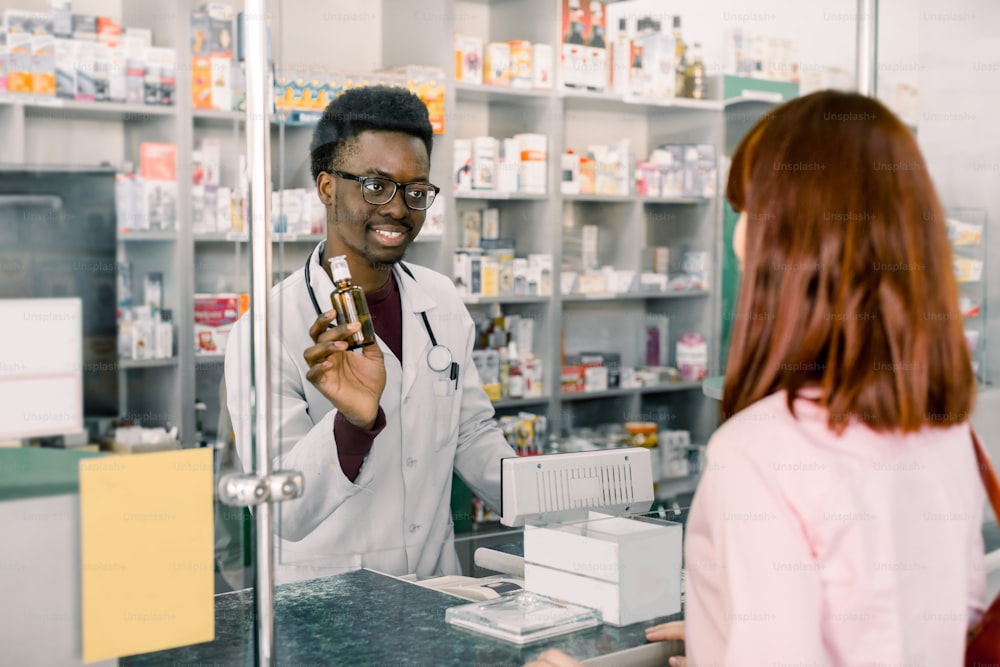 medicine, pharmaceutics, health care and people concept - happy young African man pharmacist giving drug to young pretty woman customer at drugstore.