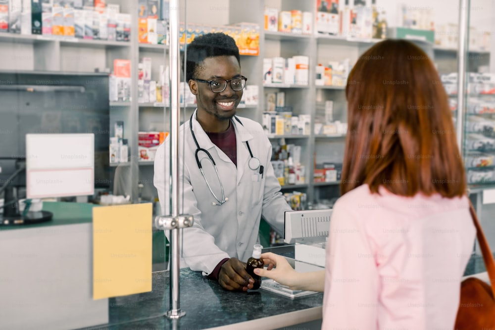 smiling male Afrcian pharmacist giving medicine to young Caucasian woman in drugstore.