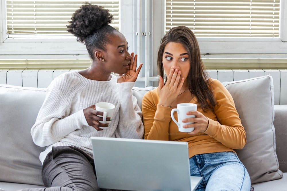 Diverse female friends sharing secrets. Two women gossiping at home. Excited emotional girl whispering to her friend ear while sitting in living-room