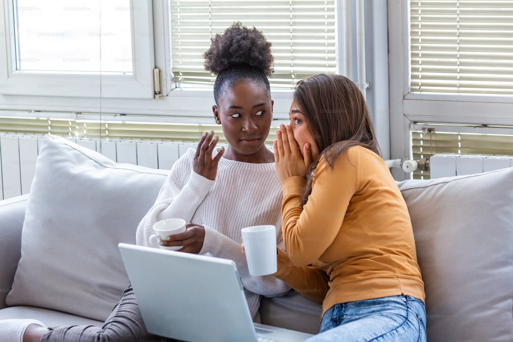 Diverse female friends sharing secrets. Two women gossiping at home. Excited emotional girl whispering to her friend ear while sitting in living-room