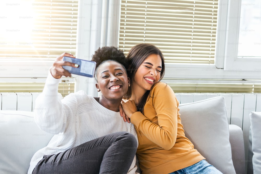 Laughing woman friends hugging each other on sofa while taking selfie photo on smart phone. Lovable caucasian and African American girls expressing positive emotions to camera.