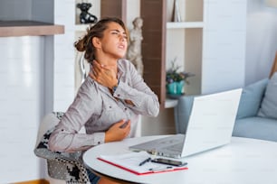 Portrait of young stressed woman sitting at home office desk in front of laptop, touching aching back with pained expression, suffering from backache after working on pc