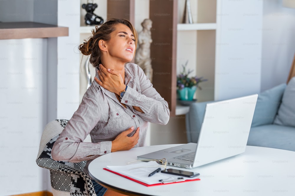 Portrait of young stressed woman sitting at home office desk in front of laptop, touching aching back with pained expression, suffering from backache after working on pc