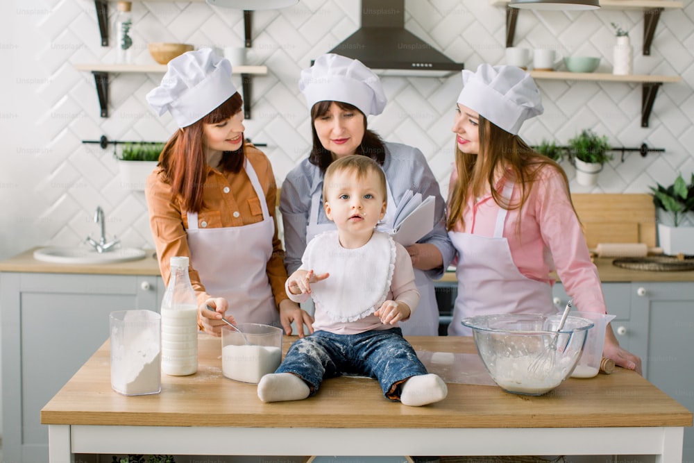 Little baby girl is sitting on the wooden table at kitchen while her mother, aunt and grandmother read the book with recipes on the background. Happy women in white aprons baking together