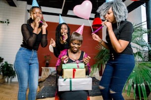 A beautiful smiling African girl opens a present at her birthday party. Happy african girls in party hats and with blowing horns are standing around birthday girl and smiling
