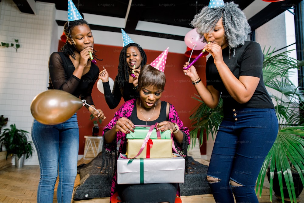 Ein wunderschönes lächelndes afrikanisches Mädchen öffnet ein Geschenk auf ihrer Geburtstagsfeier. Fröhliche afrikanische Mädchen in Partyhüten und mit Blashörnern stehen um Geburtstagskind herum und lächeln